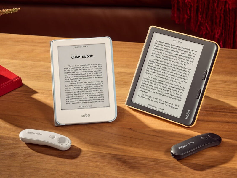 A white Kobo Clara Colour and black Kobo Libra Colour are propped up on a table in their SleepCovers with built-in stands, alongside two Kobo Remotes, one white and one black.