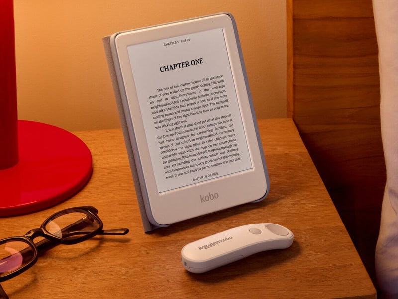 A white Kobo Clara Colour is propped up on table in its SleepCover with built-in stand, alongside a white Kobo Remote and a pair of eyeglasses.